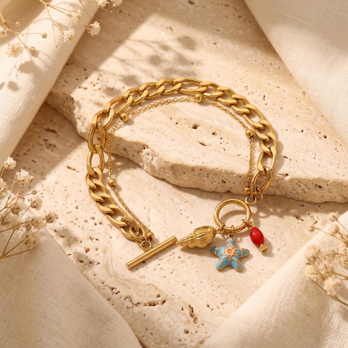 Unclasped gold-tone curb-chain bracelet with shell, blue starfish, and red charms on a textured stone surface.