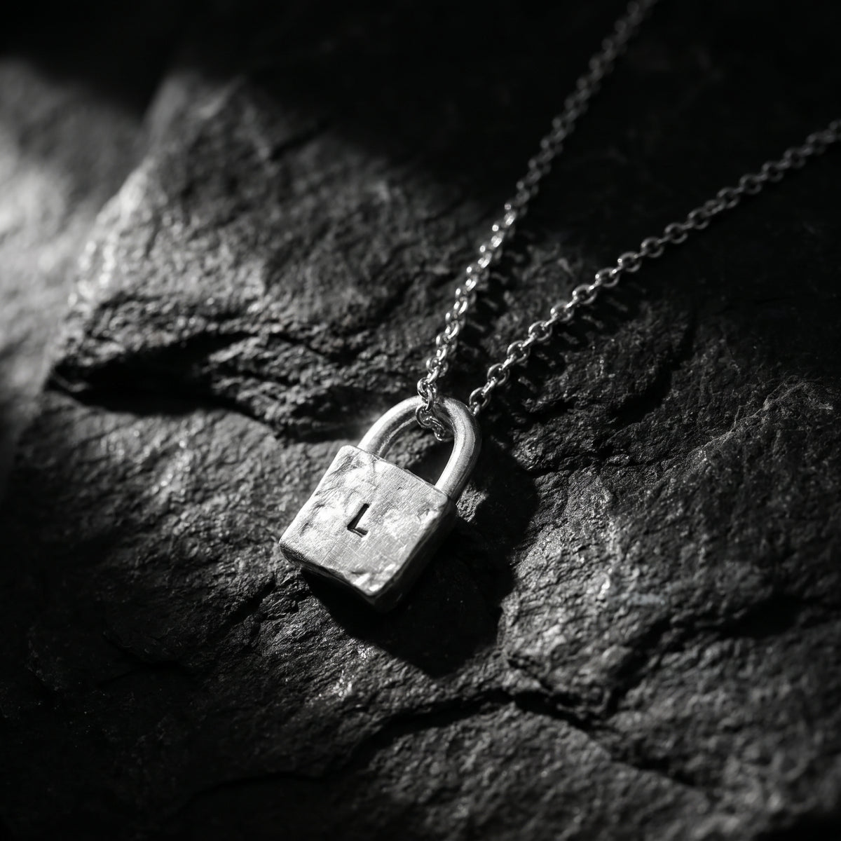 Angled close-up of the textured silver initial L lock pendant resting on dark slate rock.