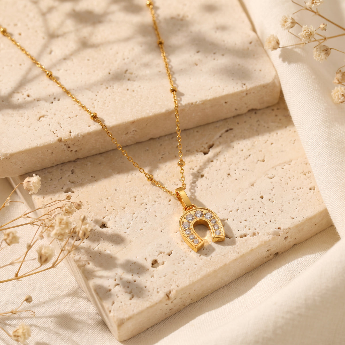Gold-tone horseshoe necklace with clear stones laid across textured stone slabs beside dried flowers.