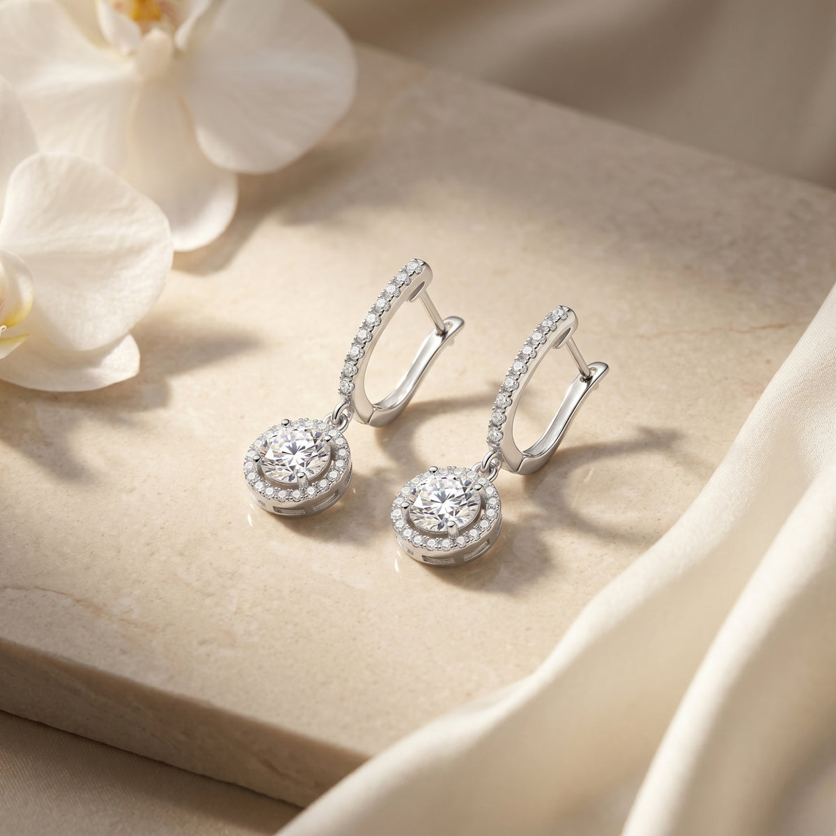 Pair of silver round moissanite drop earrings on a stone surface with white orchids.