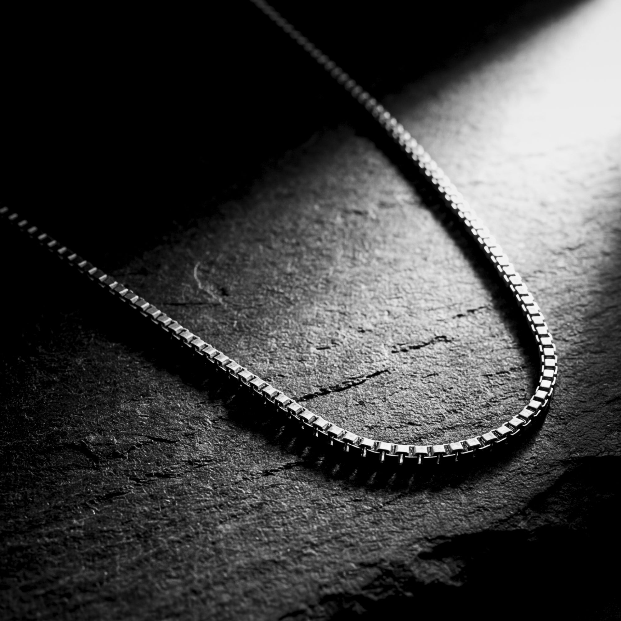 Close-up of polished silver stainless steel box chain links on a dark slate surface.