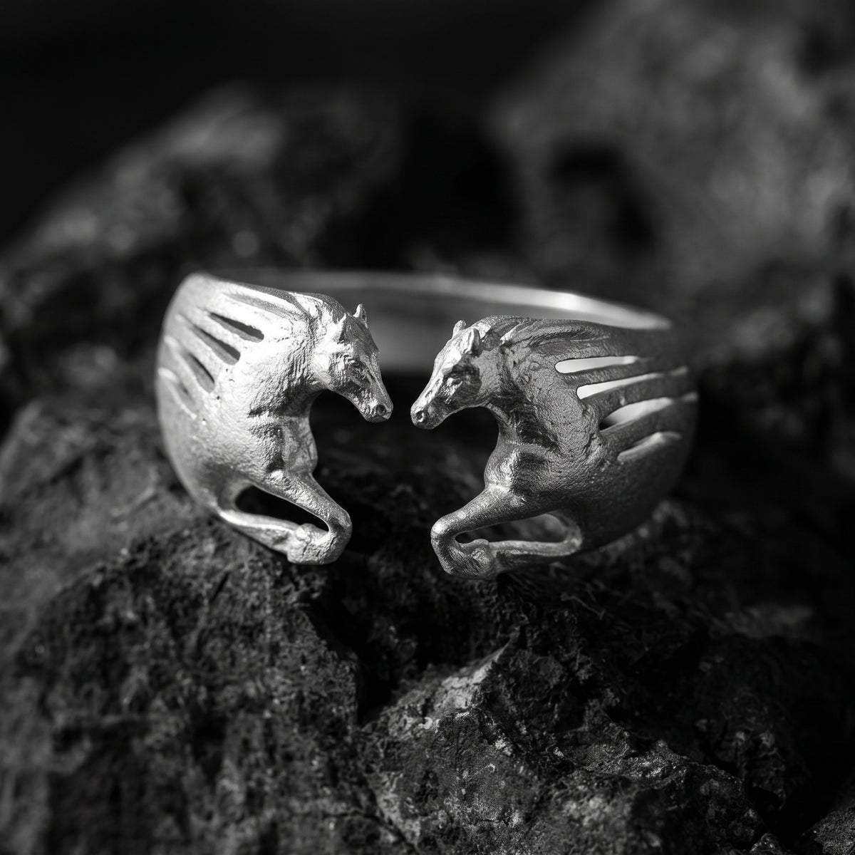 Front close-up of the silver open ring on dark rock, showing two sculpted horses facing inward across the gap.