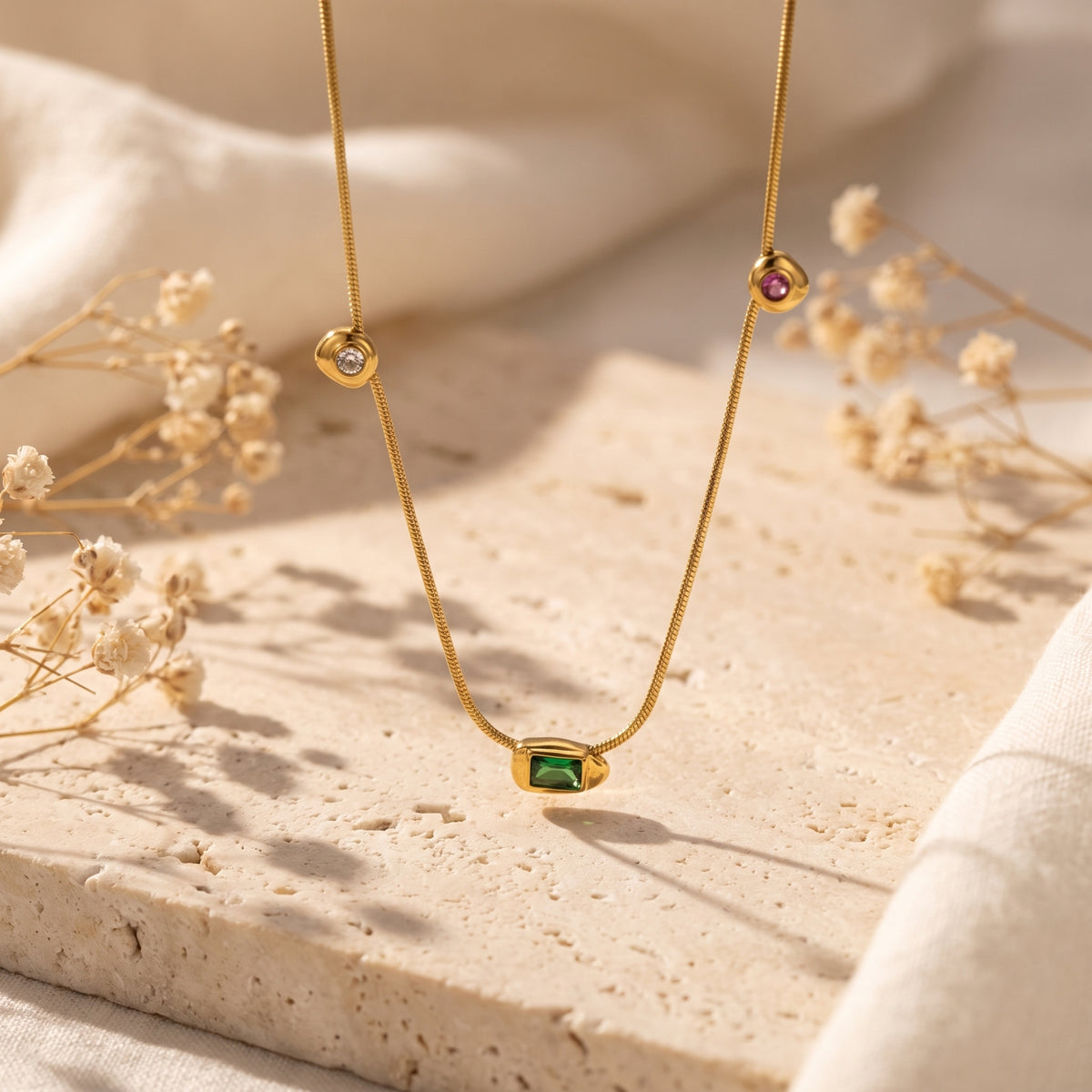 Gold station necklace suspended over a textured stone surface, with a green center pendant and round red and clear stations.