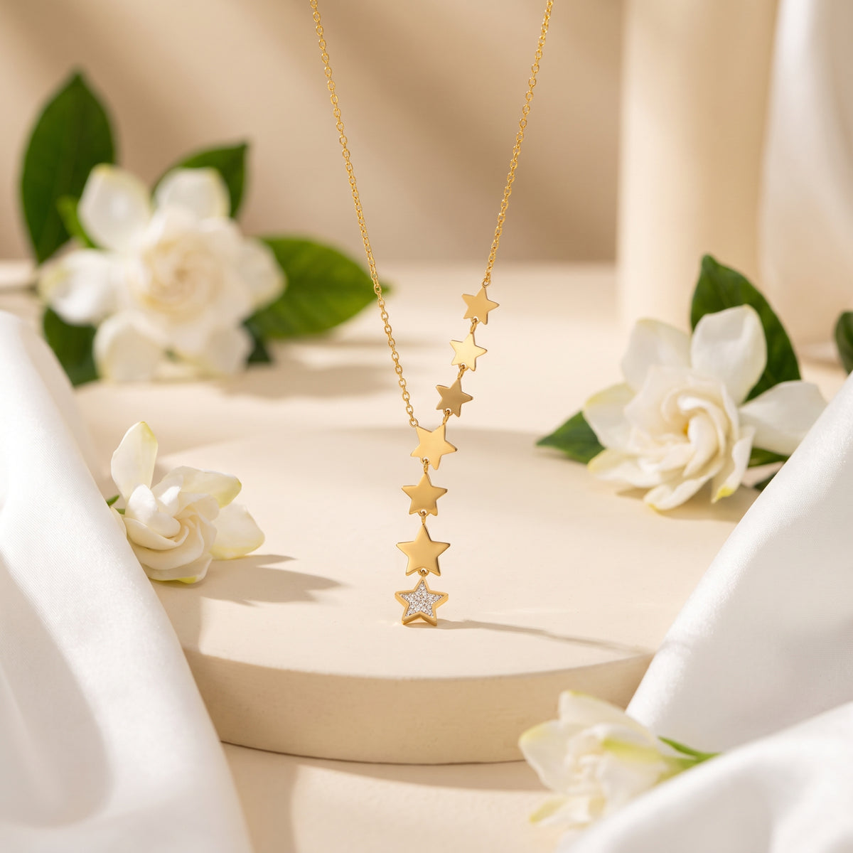 Gold-tone star lariat necklace hanging over a round display pedestal surrounded by white flowers.