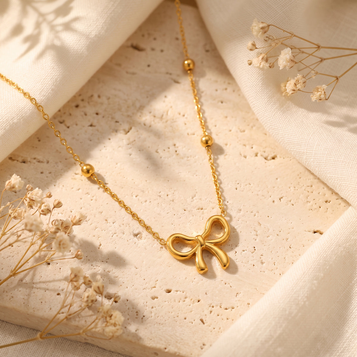 Gold-tone bow station necklace arranged on a light beige textured surface with dried flowers and white fabric.