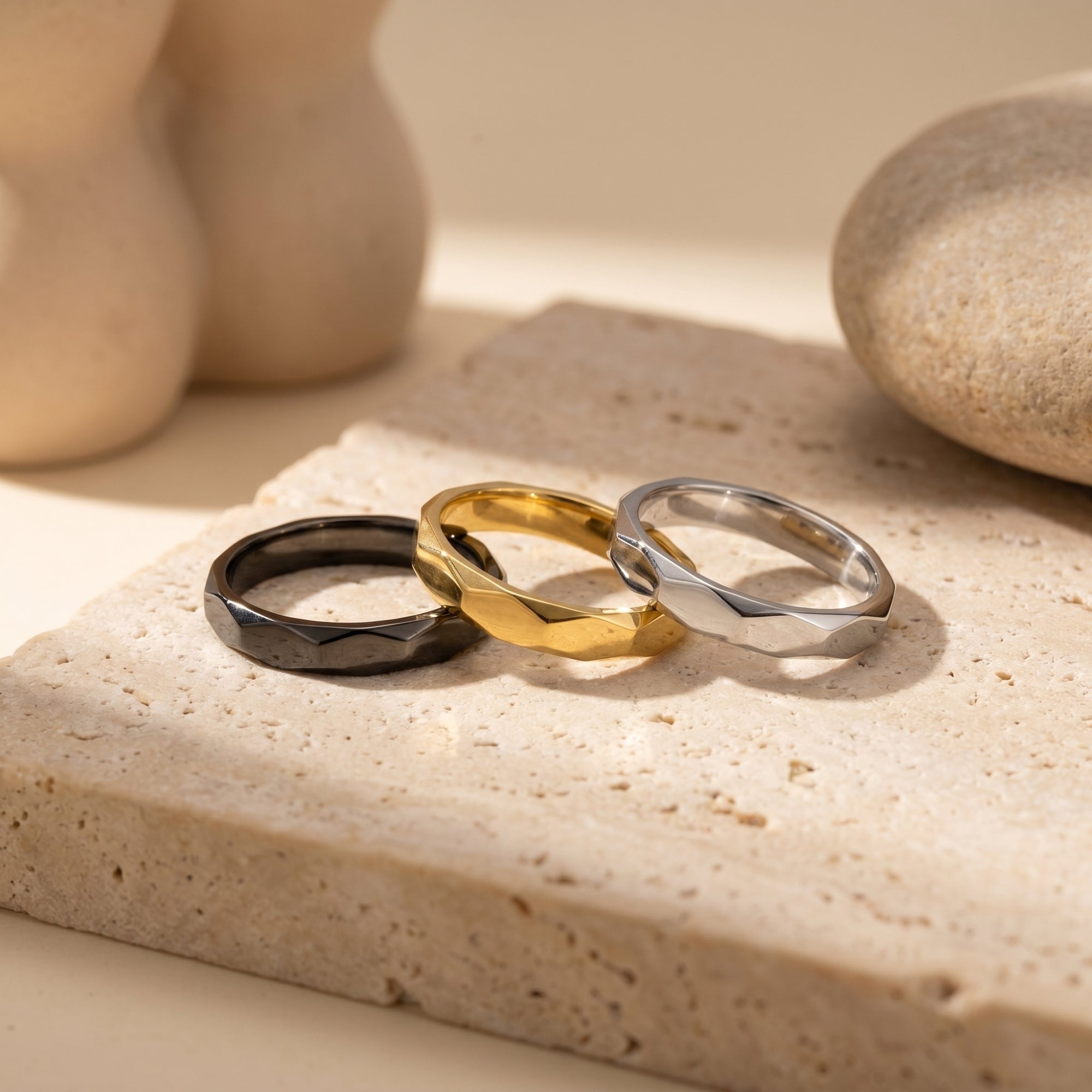 Macro view of black, gold, and silver faceted stainless steel bands resting on a textured stone surface.