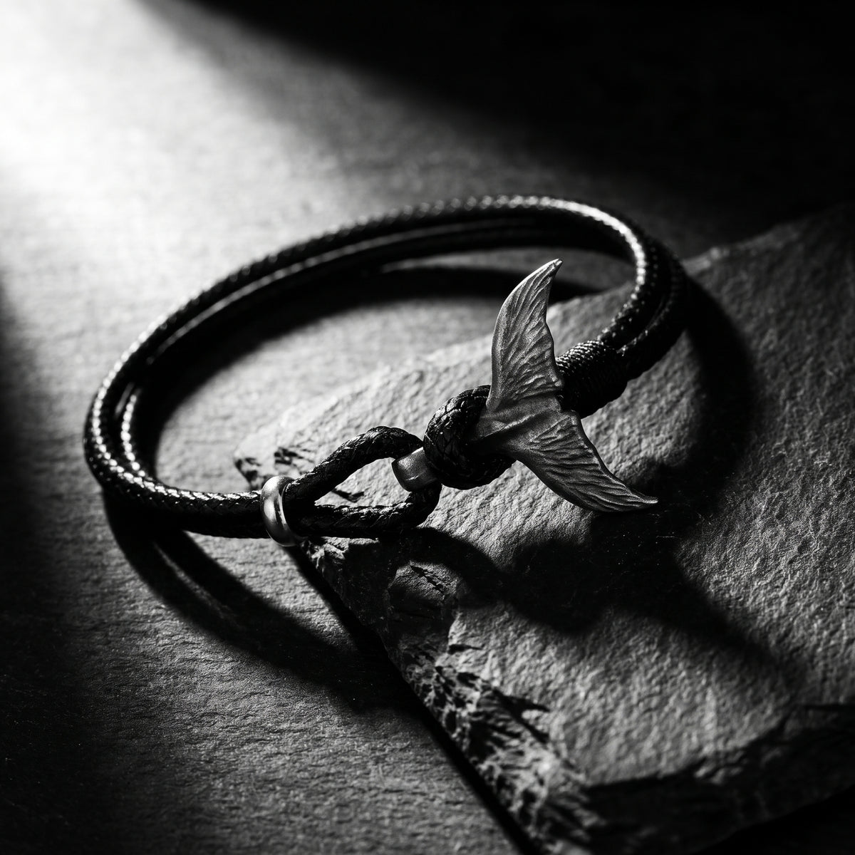 Angled close-up of the black cord bracelet and silver whale tail clasp displayed on a slate surface.