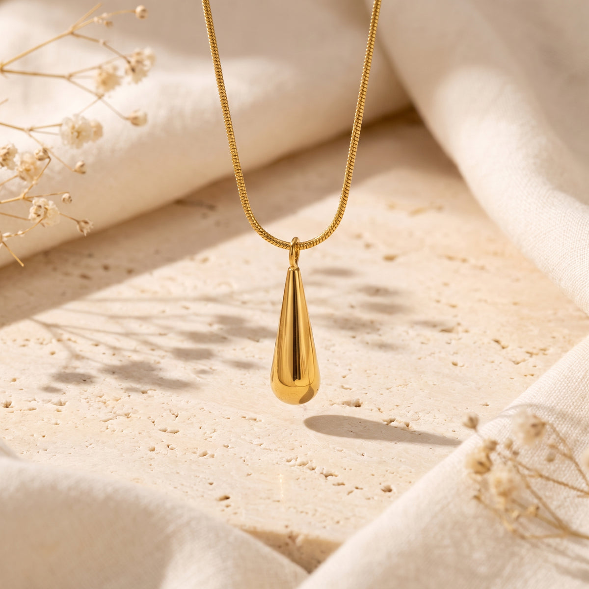 Gold-tone teardrop pendant hanging from a snake chain over a textured stone surface with dried flowers.