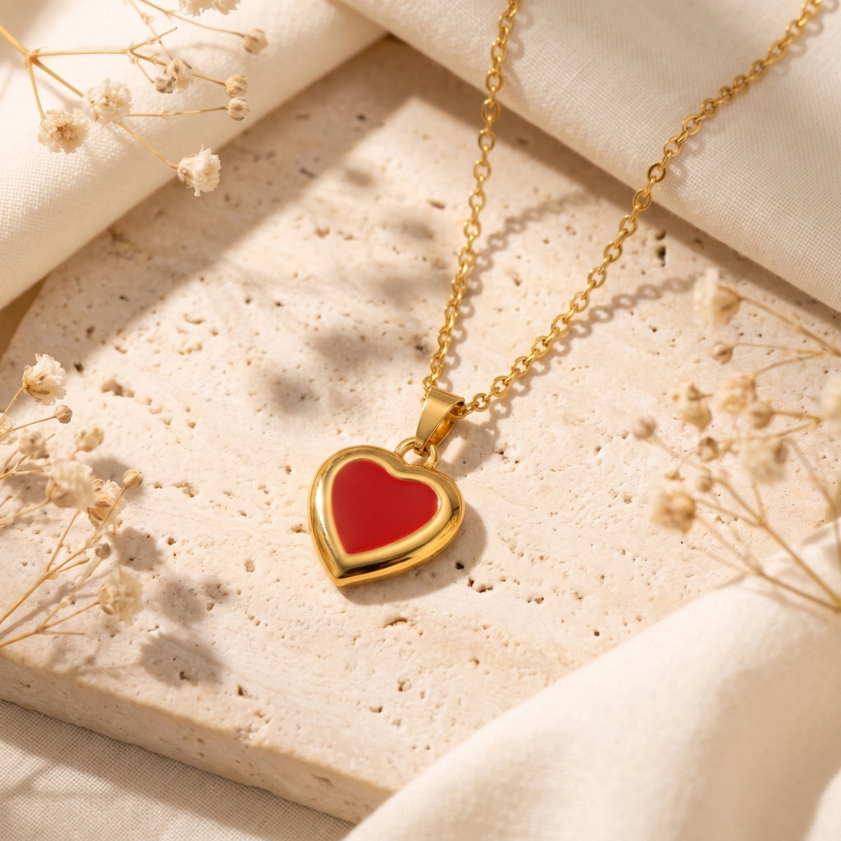 Red enamel heart pendant necklace arranged flat on a pale stone slab with dried flowers.