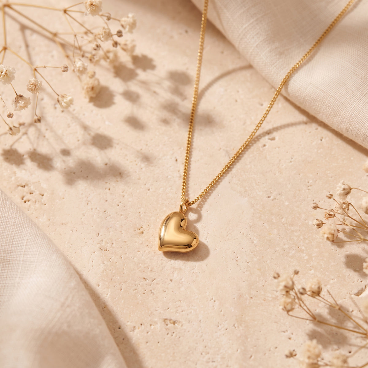 Gold-tone puffed heart pendant necklace arranged on a beige stone surface with linen fabric and dried flowers.