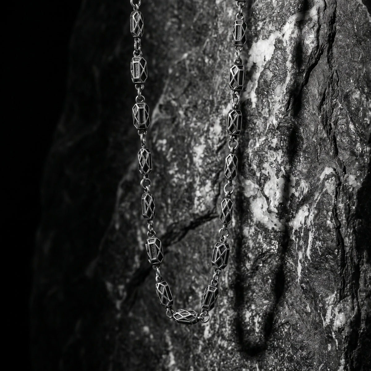Silver geometric link necklace hanging vertically against a dark, rough stone surface.