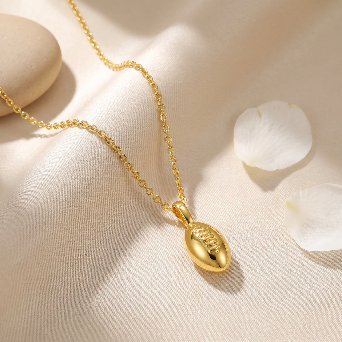 Flat-lay of a gold-tone football pendant necklace on cream fabric beside white petals and a pale stone.