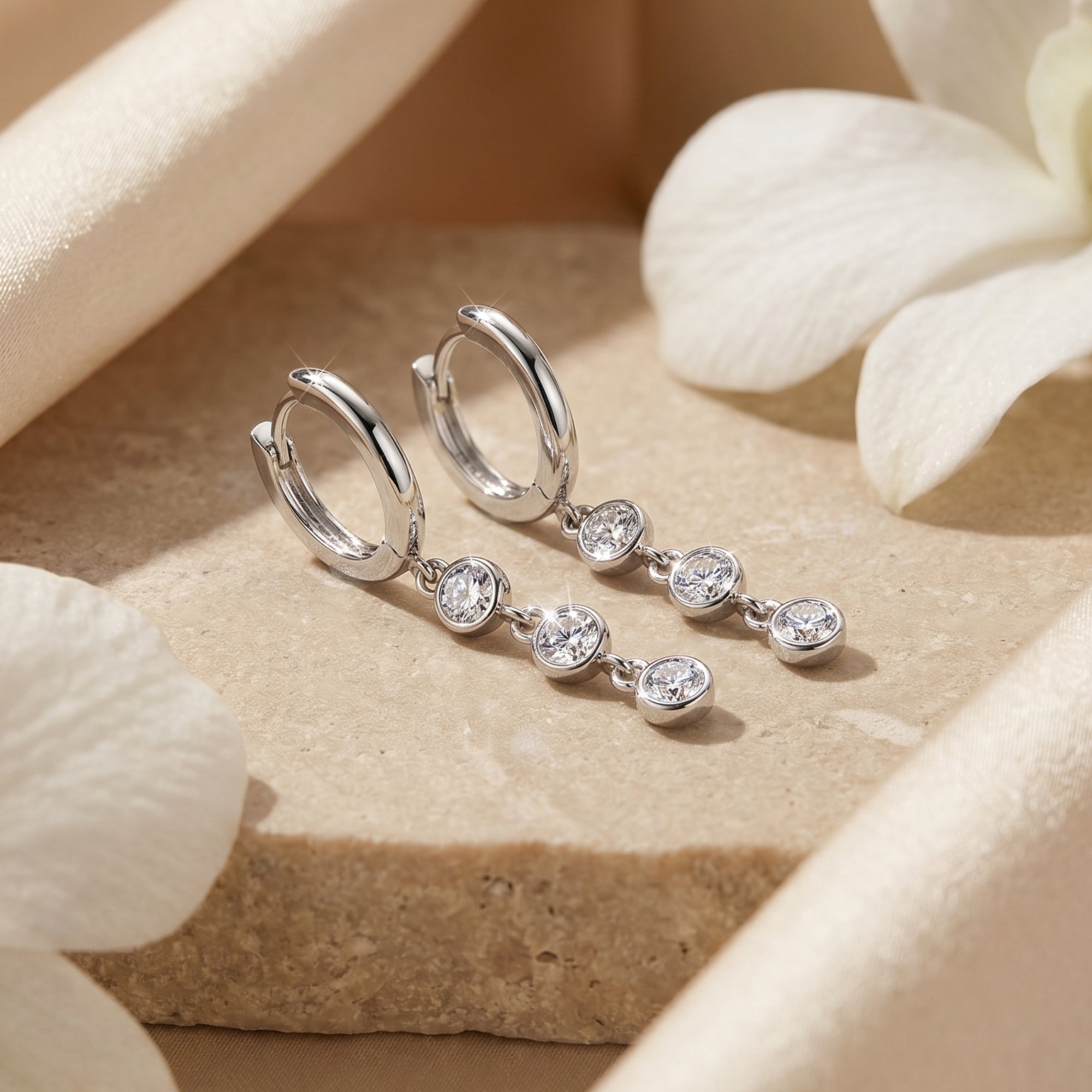 Silver moissanite drop earrings with three round stones on a textured stone surface with white petals.