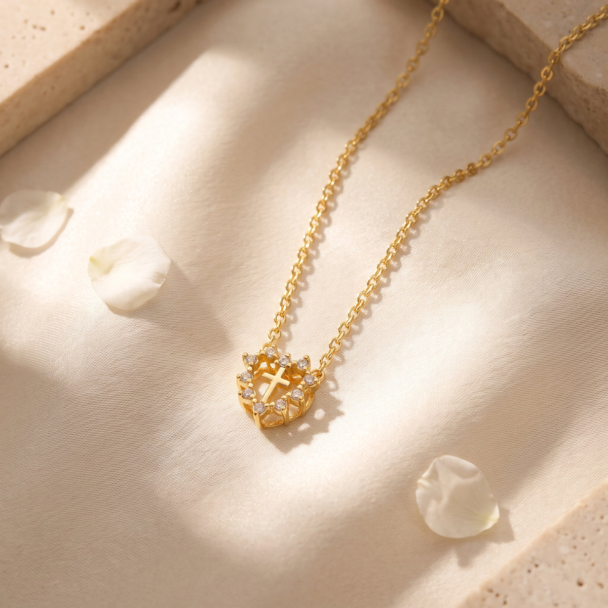 Gold-tone heart halo cross necklace laid on light fabric with white petals and stone blocks.
