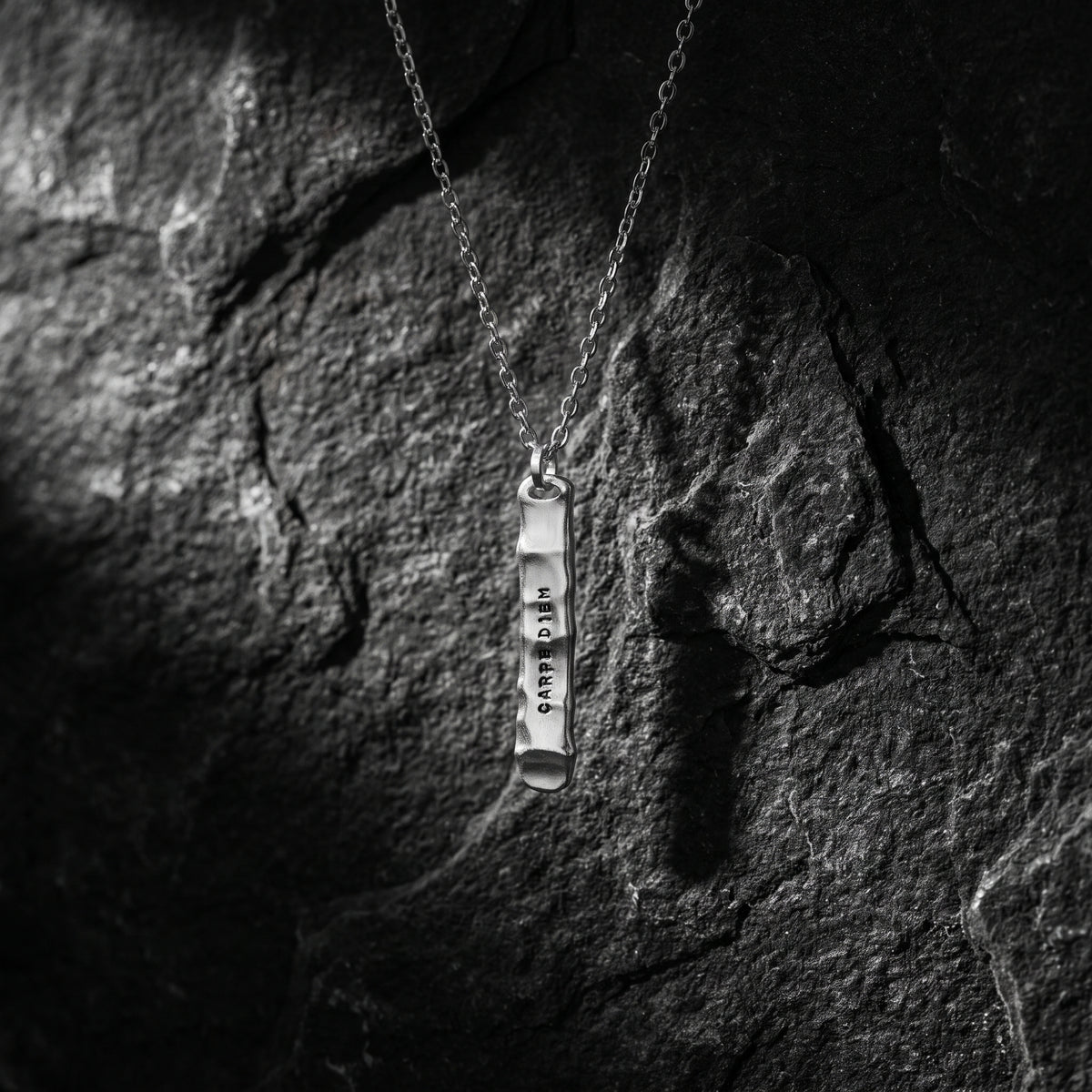 Silver bar pendant with CARPE DIEM text hanging straight against a dark stone surface.