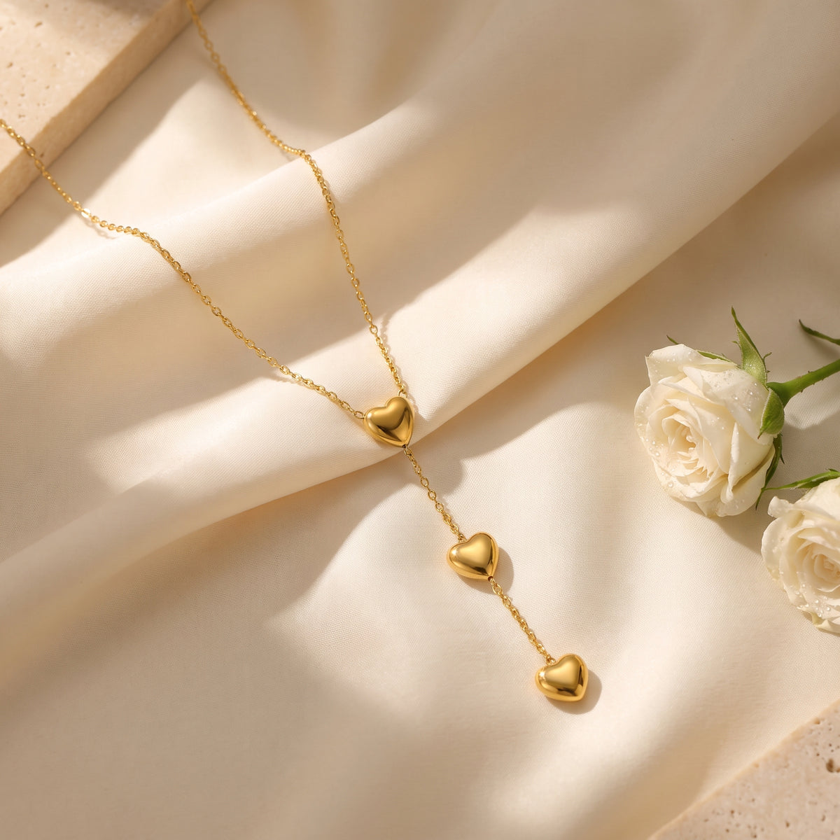 Gold-tone triple heart lariat necklace arranged on draped cream fabric beside white roses