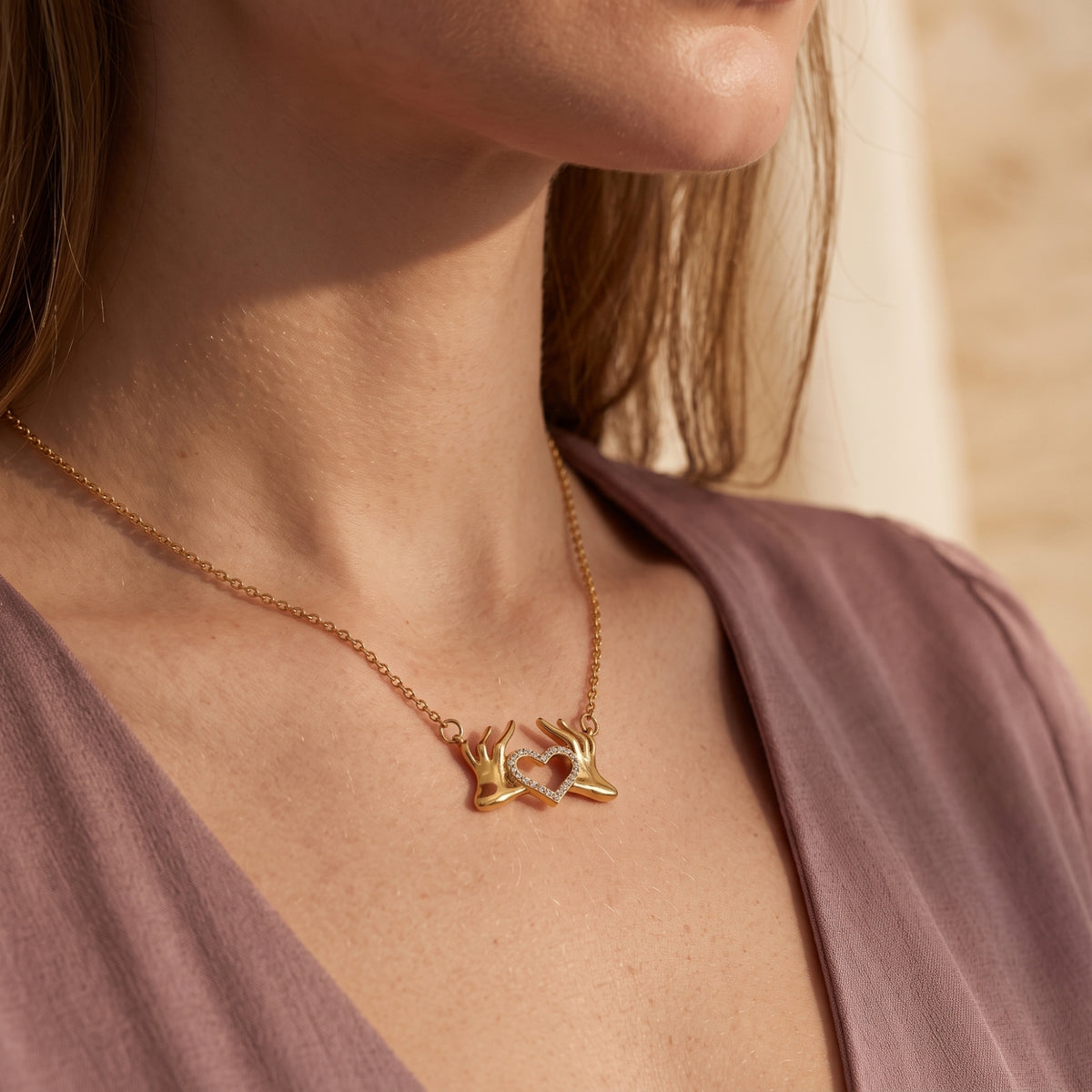 Close-up of the gold-tone heart hands pendant necklace worn with a mauve V-neck garment.