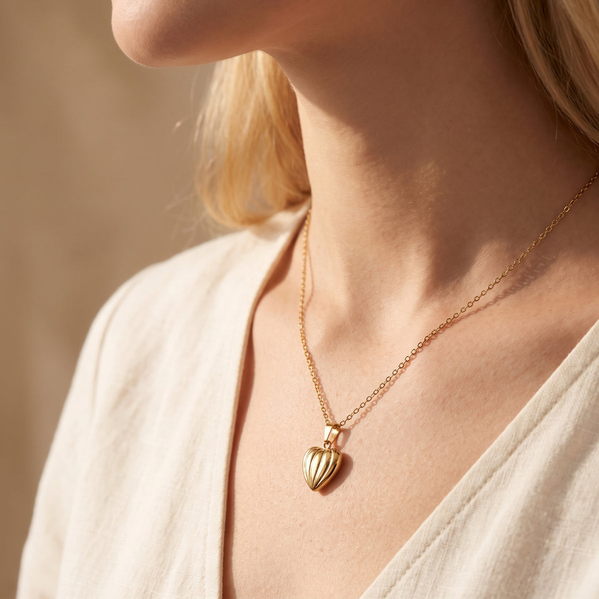 Close-up profile view of a woman wearing a gold-tone ribbed heart pendant necklace over a light-colored open-neck top.
