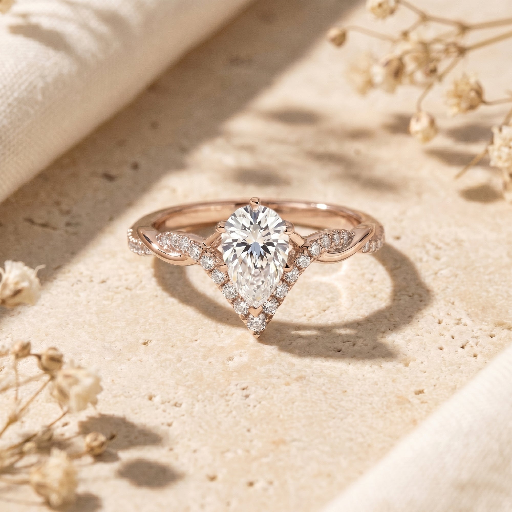 Rose gold pear-cut moissanite promise ring with a chevron halo and twisted band on a stone surface.