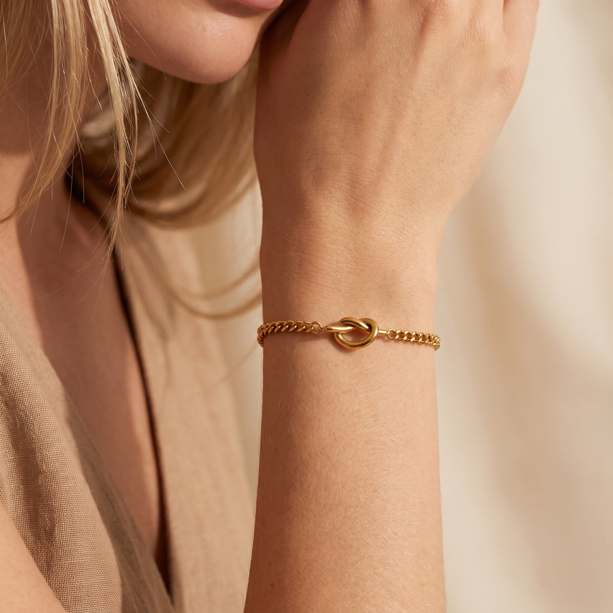 Gold-tone knot link bracelet worn on a wrist beside a neutral-toned outfit.