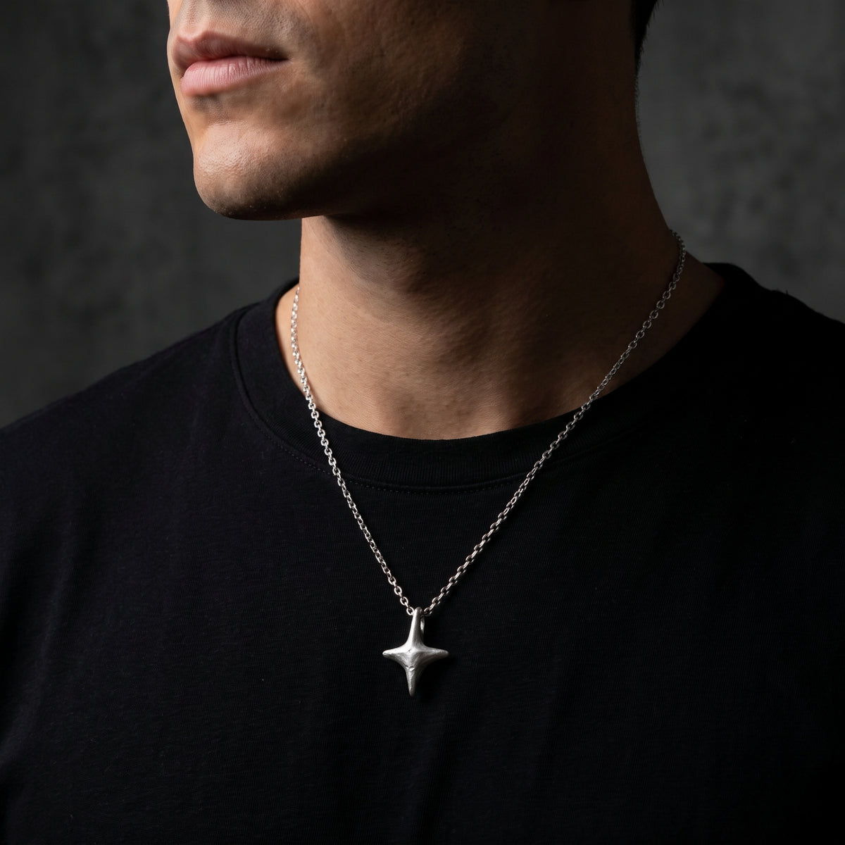 Close view of a man wearing a silver four-point starburst pendant over a black crewneck shirt.