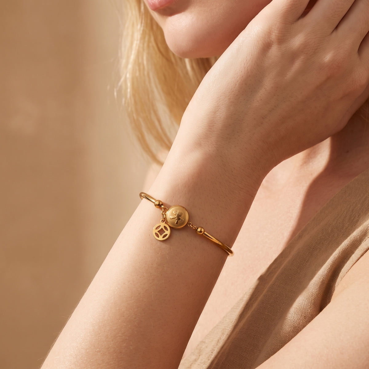 Model wearing a gold-tone station bracelet on her wrist beside her face, with a round medallion and dangling openwork charm.