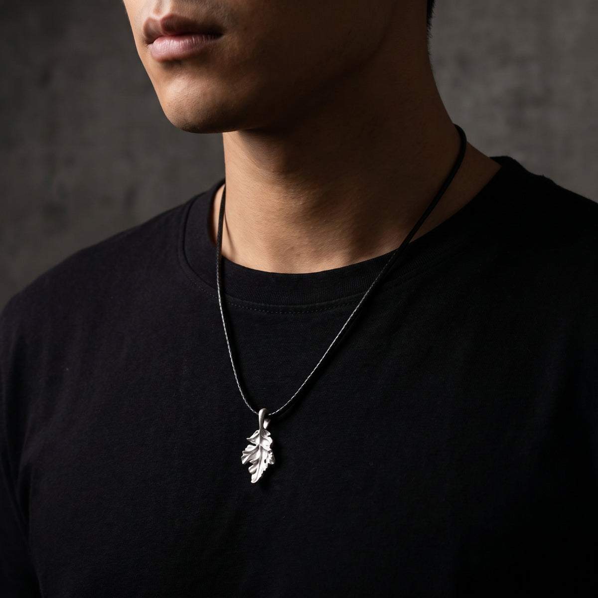 Silver oak leaf pendant worn on a black cord against a black shirt, shown from a side angle.