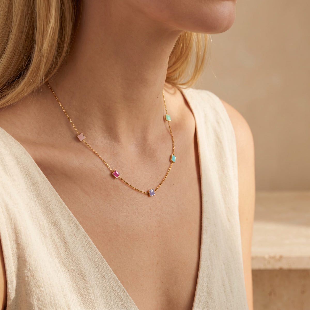 Blonde model wearing a gold-tone enamel square station necklace over a cream V-neck top.