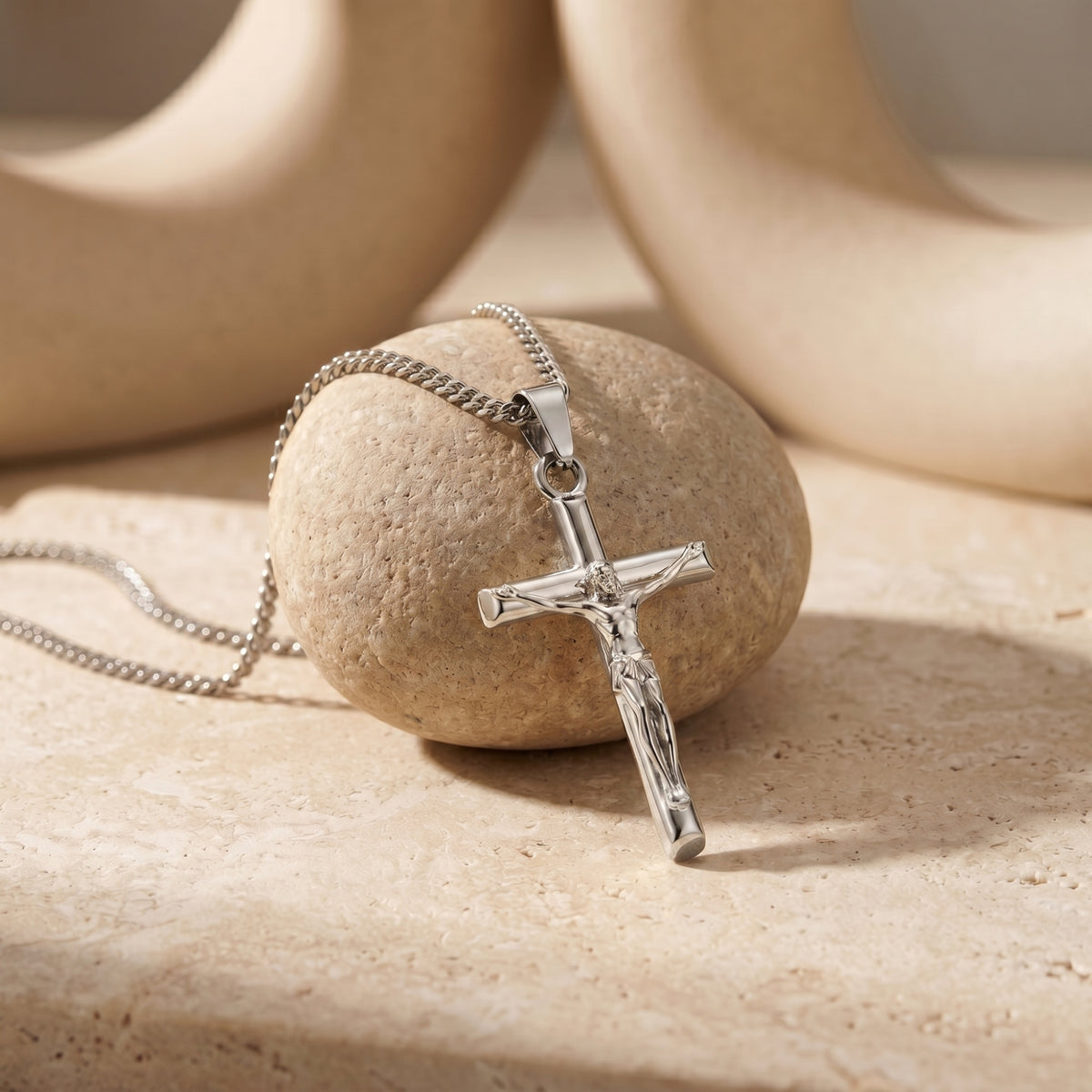 Silver crucifix pendant and curb chain draped over a textured round stone on a beige surface.