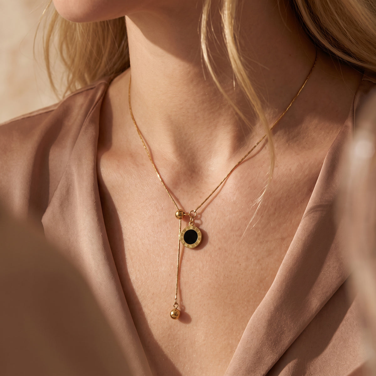 Gold-tone lariat necklace with black Roman numeral disc pendant worn on a model in a taupe open-front garment.
