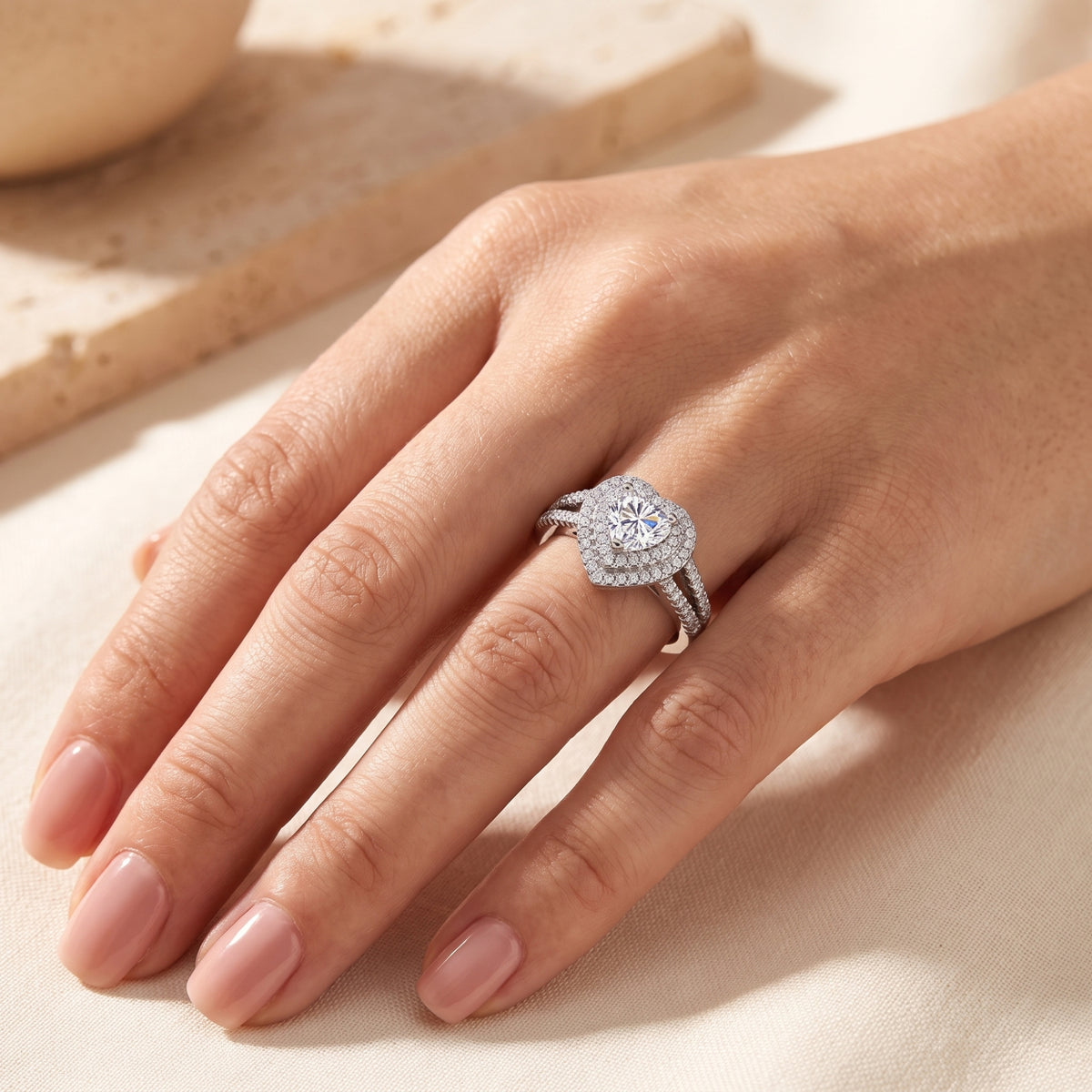 Silver heart halo ring on a hand resting over textured beige cloth, showing the central stone and shank.