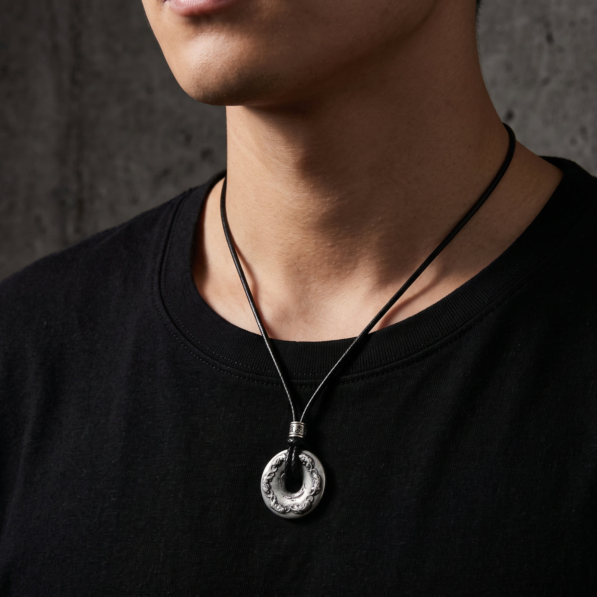 Man wearing the carved silver disc necklace, positioned mid-chest over a black crewneck shirt.