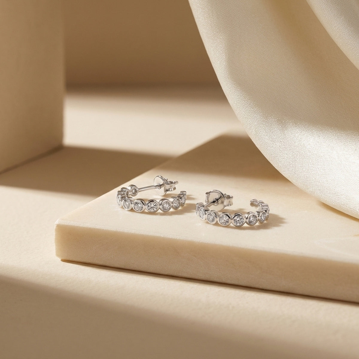 Silver-tone bezel-set moissanite hoop earrings displayed front-facing on a beige block beside draped fabric.