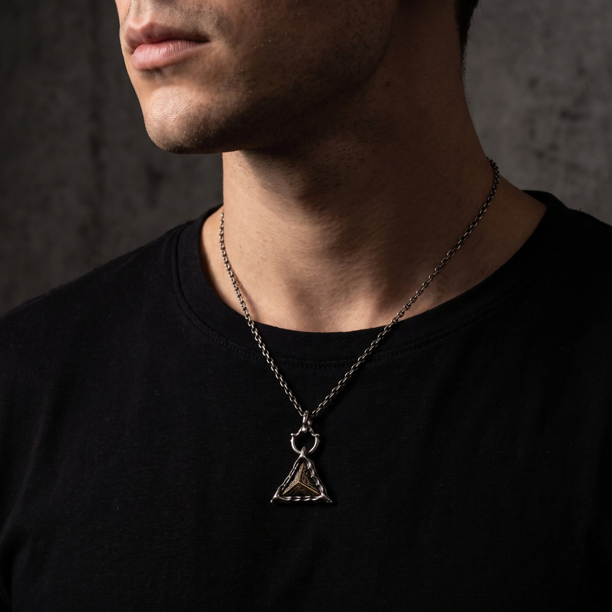 Close-up side view of a man wearing the silver triangular pendant necklace over a black shirt.