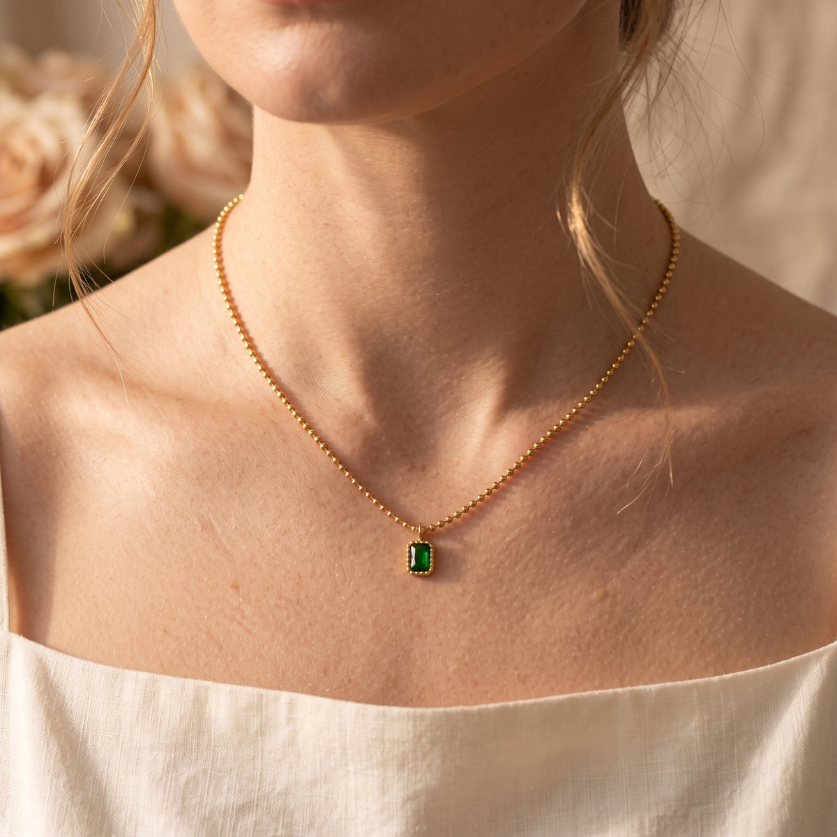 Frontal view of a model wearing the gold-tone ball chain necklace with a green emerald-cut pendant.