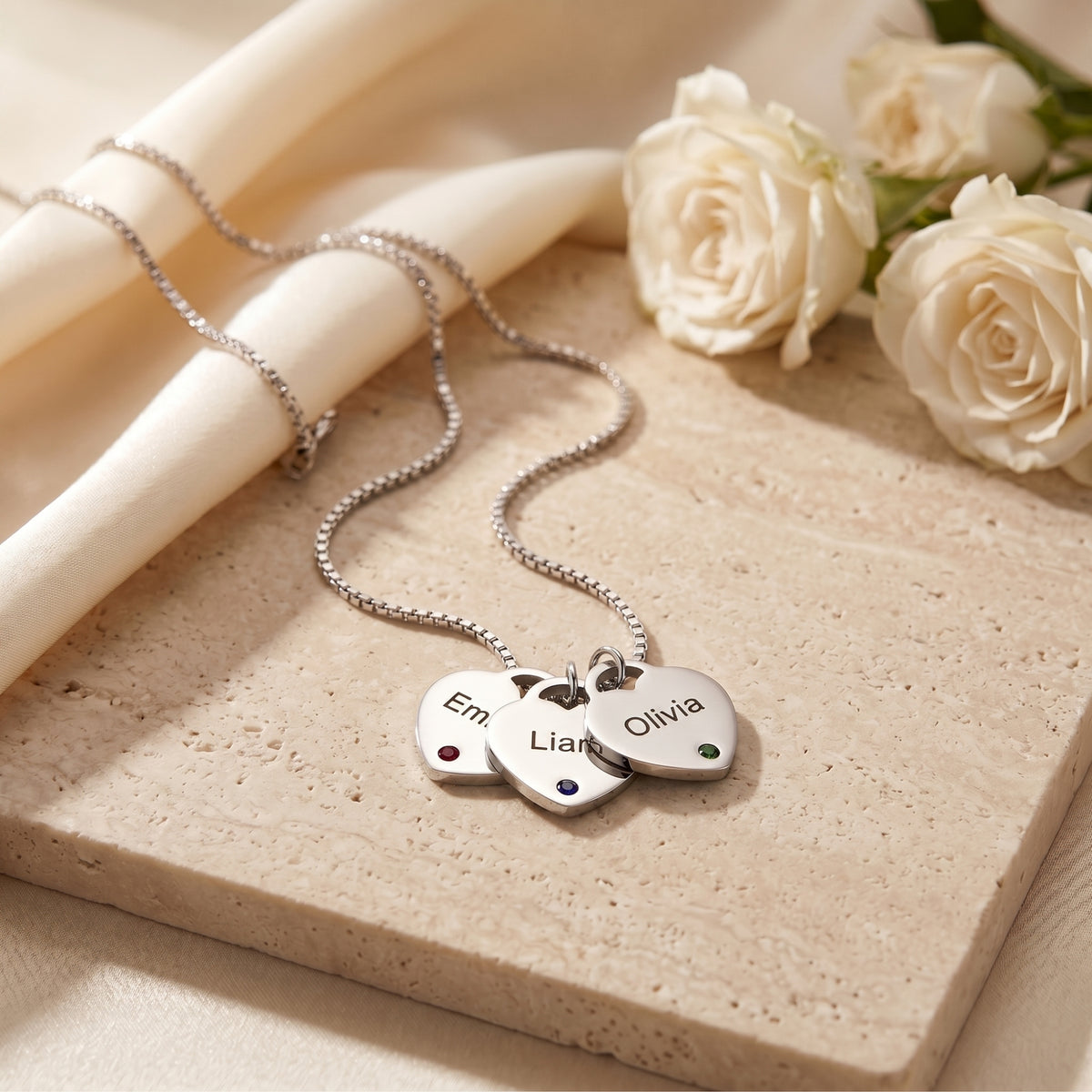 Silver Sybilla necklace with three engraved heart pendants and birthstones on a stone slab with white roses.