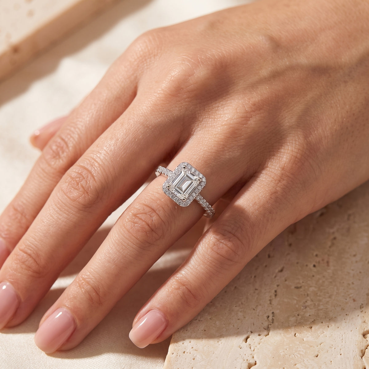 Angled view of a silver-toned emerald-cut halo ring worn on a hand over a cream stone background.