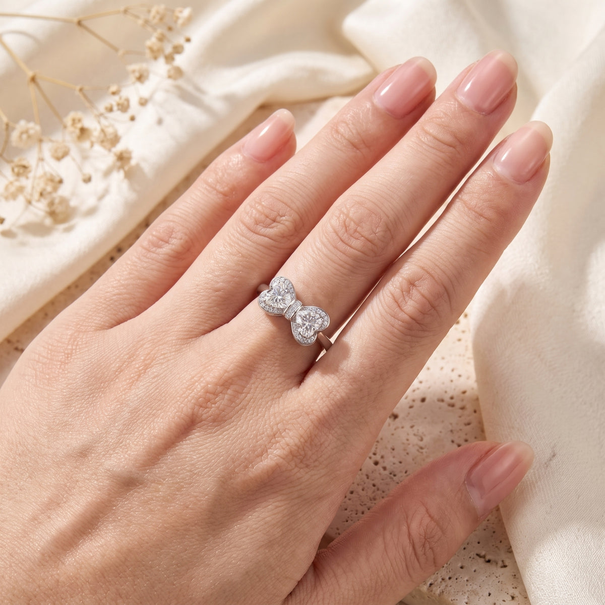 Top view of a hand wearing a bow ring with twin clear heart-shaped stones on the ring finger over draped cloth.