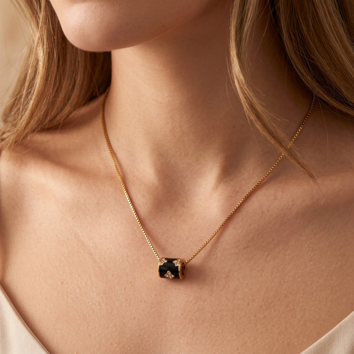 Front-facing model wearing a gold-tone box chain with a black barrel pendant below the collarbone.