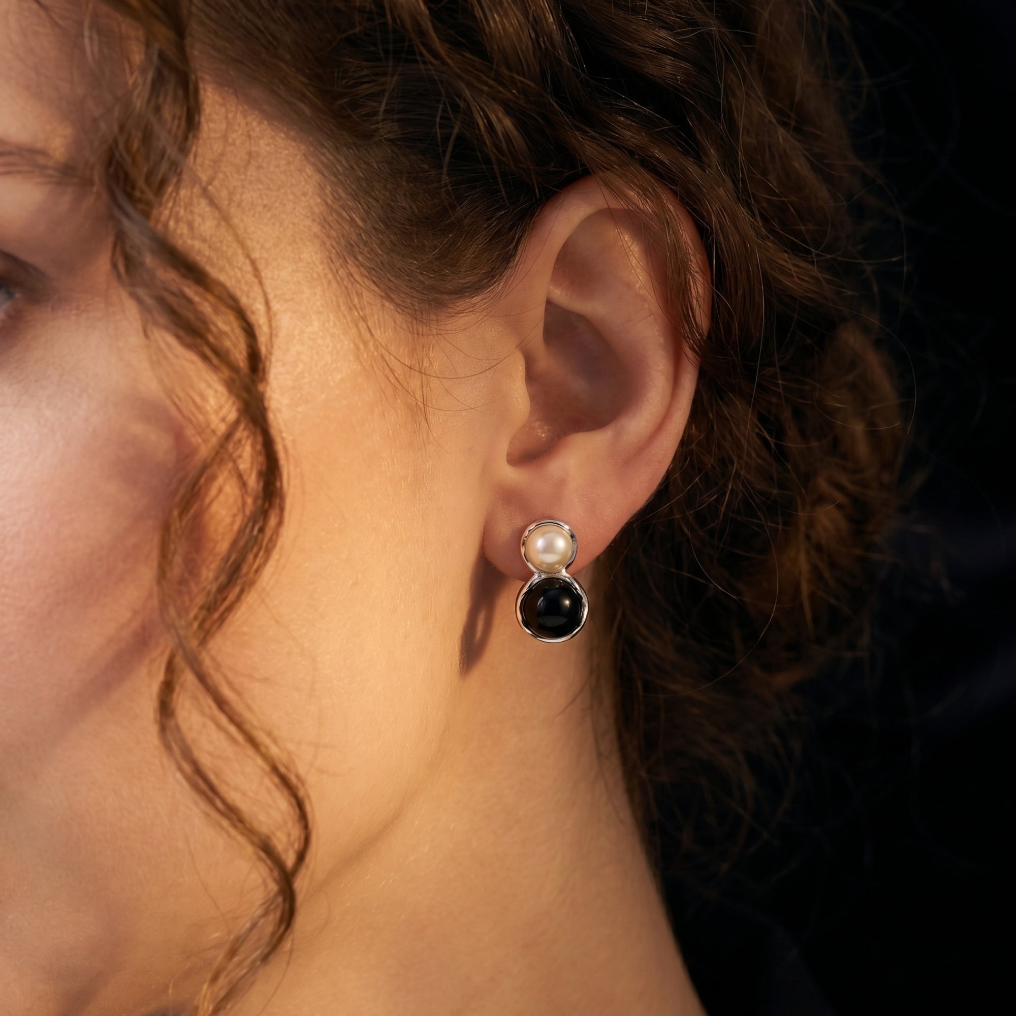 Close-up of a silver drop earring with a round white pearl and black onyx cabochon worn on a model's ear.