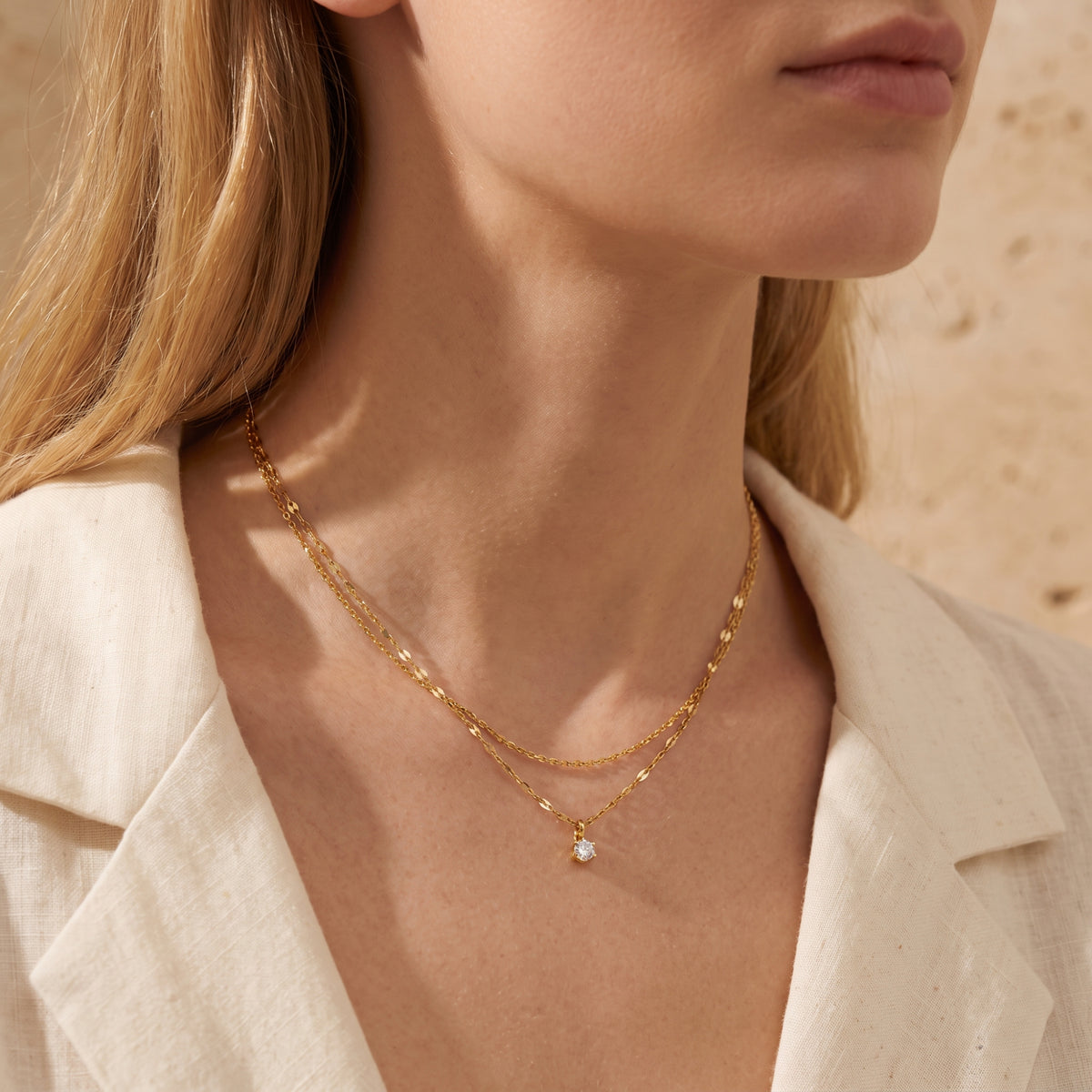 Layered gold-tone solitaire necklace shown on a model styled with a cream linen collared jacket.