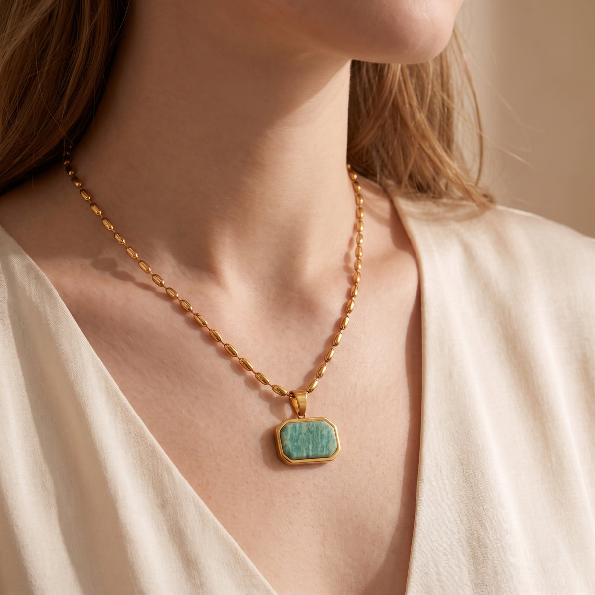 Front view of a model wearing a gold-tone bead chain necklace with a green Amazonite pendant over a cream blouse.