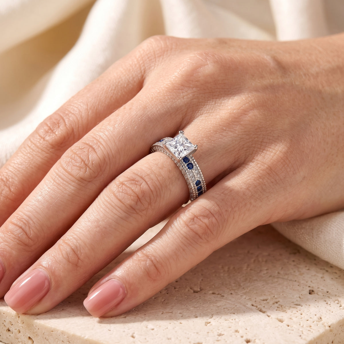 Silver princess-cut engagement ring with blue accents shown on a hand.