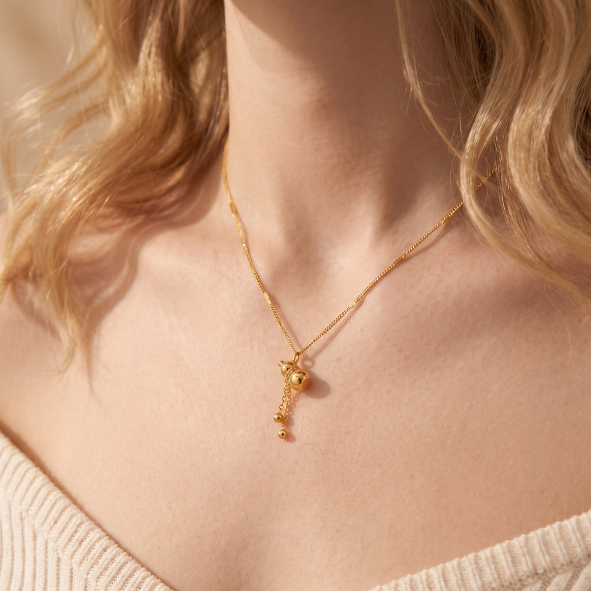 Close-up of a gold-tone gourd pendant necklace worn at a model's collarbone, with two dangling bead accents.