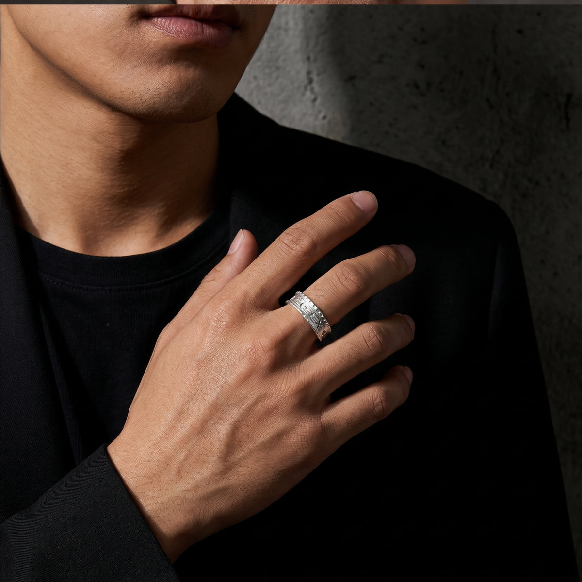 Model wearing a wide engraved silver ring on the middle finger against a black jacket.