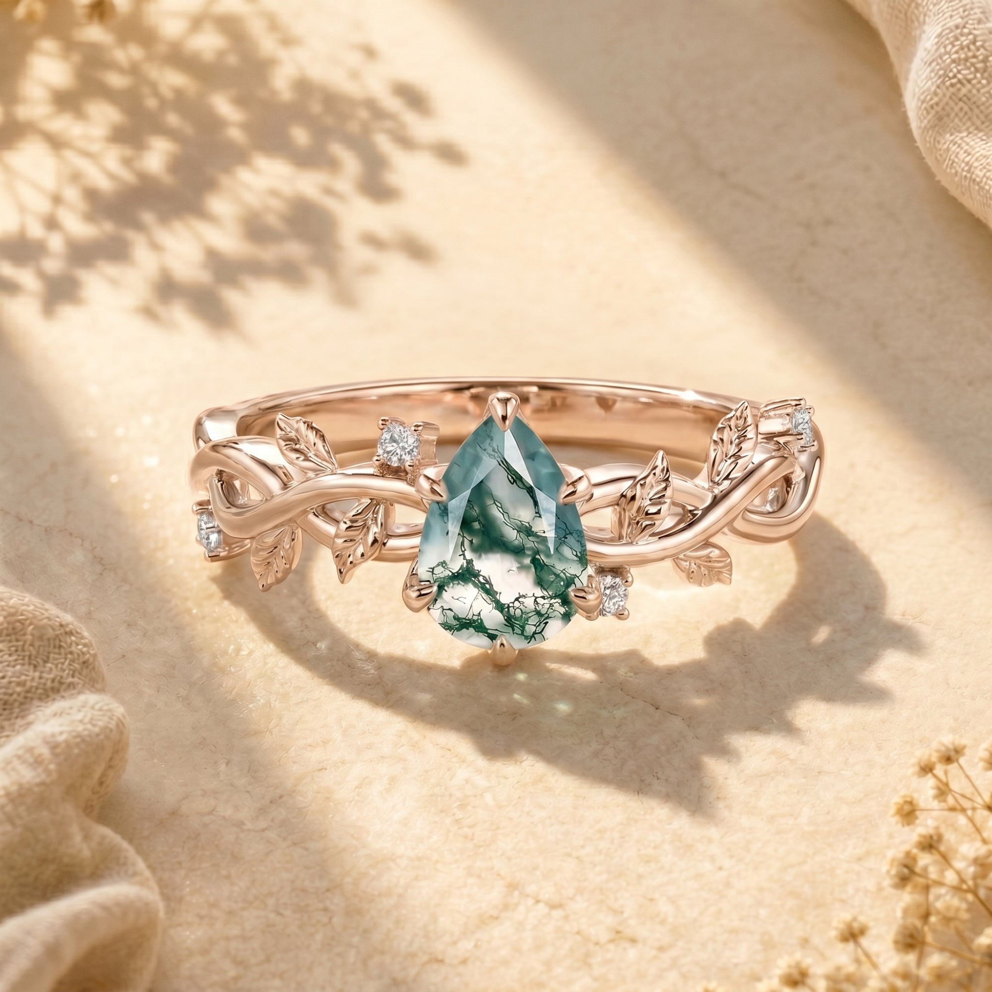 Rose gold pear-cut moss agate ring with leaf details on styled background.