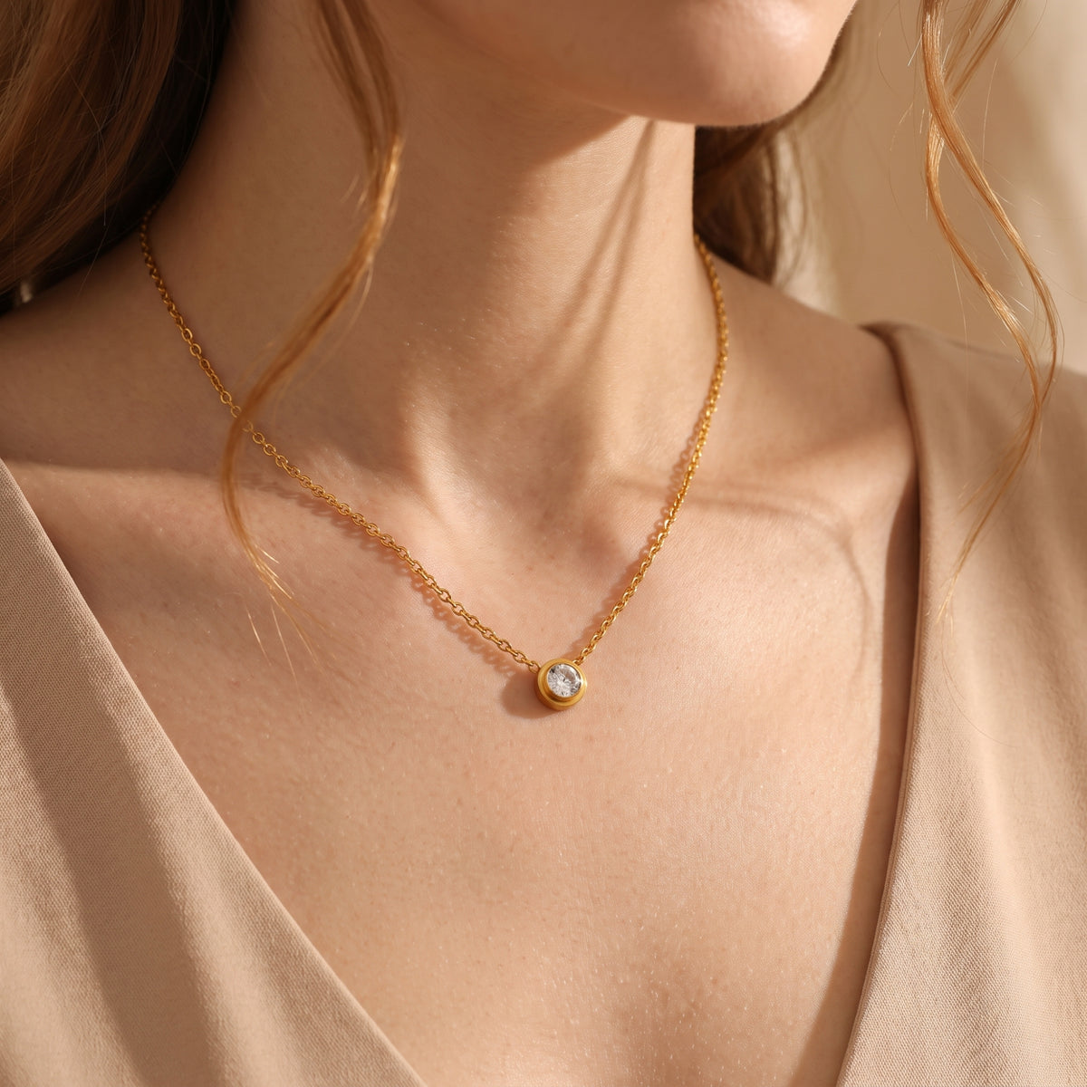Person wearing a gold-tone bezel solitaire necklace with a clear round center stone against a beige neckline.