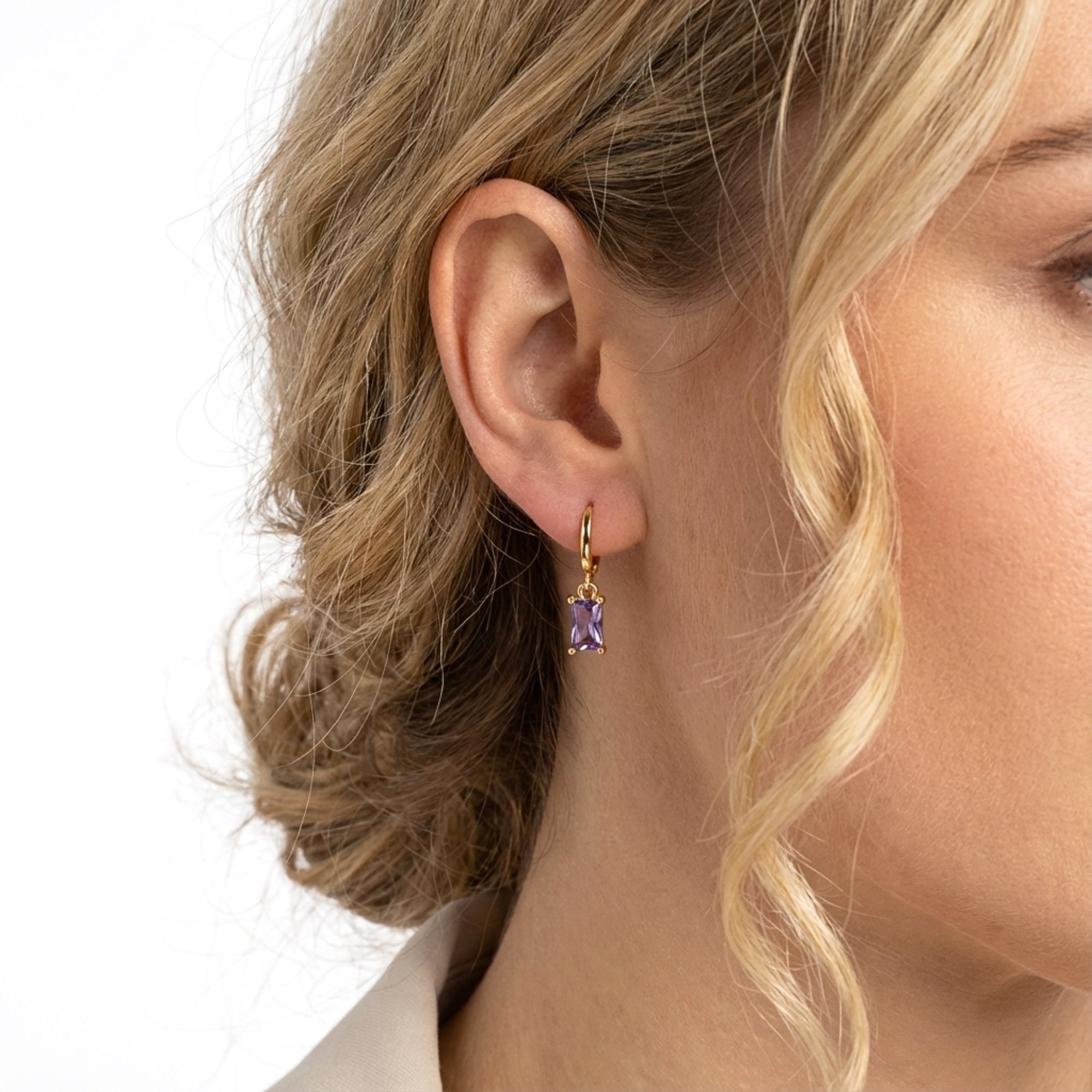 Close-up of a gold Vivienne drop earring with a purple emerald cut stone worn on a woman's ear.