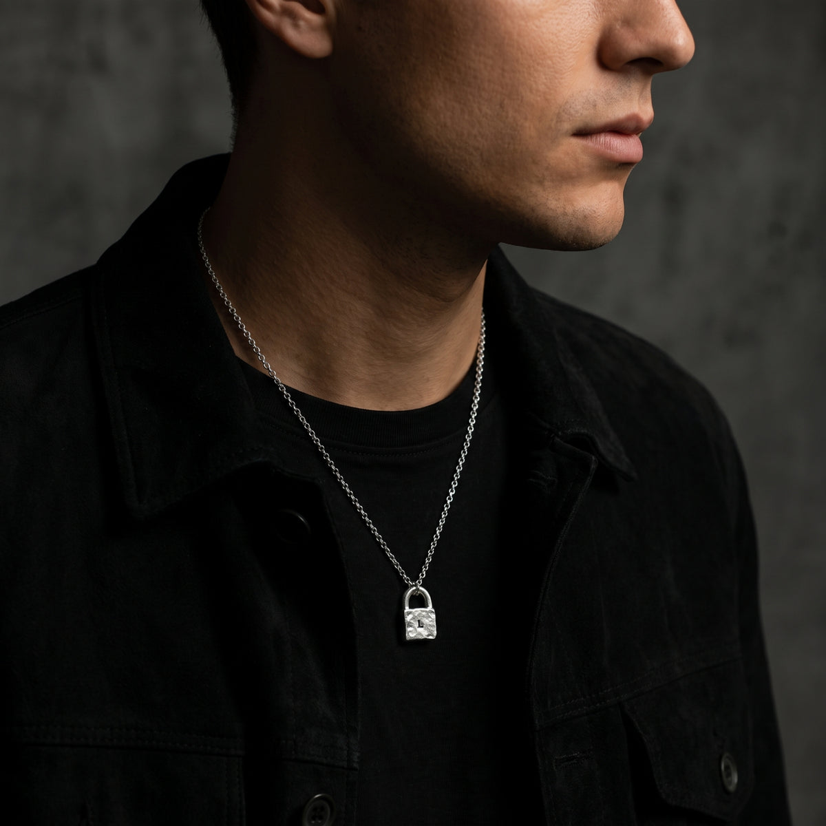 Model wearing the silver L-stamped lock necklace over a black crewneck shirt beneath a black jacket.