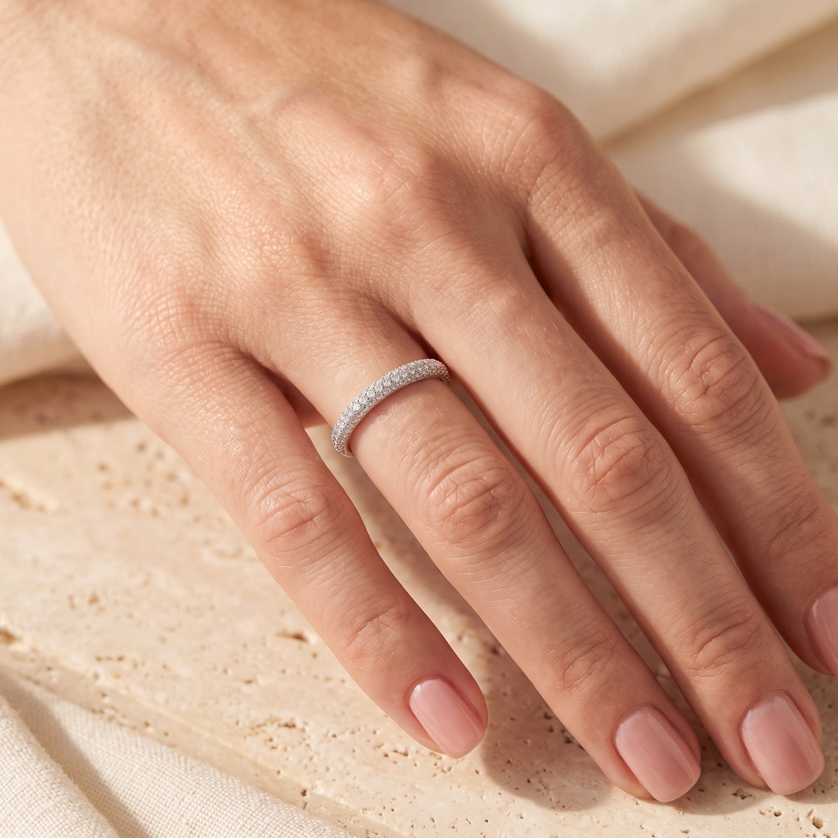 Silver micro-pavé band ring worn on a hand resting on a beige stone block.